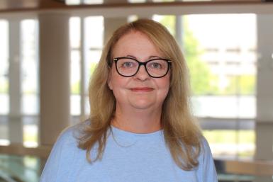 A woman with straight blond hair past her shoulders, wearing black-rimmed glasses and a light blue shirt. Behind her is a wall of windows. 