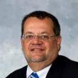 Headshot of Craig Rush, PhD. He's a white, middle-aged man with dark, short-clipped hair, and he's wearing rimless glasses, a dark suit, white collared shirt, and royal blue tie with thin, white, diagonal stripes. 