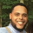 Mid-shot of Robert Bell, a Black man with a broad smile. He's wearing a light blue button-up shirt, tan suspenders, and a light gold tie, standing in front of trees. 