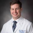 Professional headshot of Dr. Justin Huber, a white man in 30s or 40s. He has short medium-brown hair parted on his left, a kind smile, and is wearing a white lab coat, a white collared shirt, and a navy blue plaid tie. 