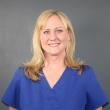 Photo of Alayne Nieto. She's a white woman with blonde hair past her shoulders. She's smiling at the camera, wearing blue scrubs, and standing in front of a gray background. 