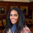 Headshot of Laneshia Conner, PhD, a Black woman in her 30s with long straight hair; she's smiling at the camera wearing salmon/blue/gray striped sweater and a blue turtle neck. In the blurred background is a maroon wall with large portraits.