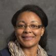 Headshot of Coretta Jenerette, a Black woman with thin rectangular glasses and her hair parted and pulled partially back. She's wearing a navy sweater with a cream colored scarf, smiling to the camera in front of a marbly gray backdrop.