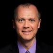 Headshot of David Gross, a middle-aged white man wearing a black suit with a light purple shirt and black and white checkered tie. He's smiling at the camera in front of a black background. 