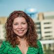 Photo of Michelle Hernandez, a middle aged woman with light skin and curly reddish-brown hair past her shoulders. She's wearing a v-neck green dress with dark blue pattern and is standing outside. 