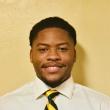 Headshot of Ellis Jackson, a young Black man wearing a white collared shirt and a tie with wide yellow and navy stripes. He's smiling at he camera and short hair, a thing mustache, a goatee. There's a beige wall beind him. 