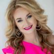 Professional headshot of Carey Beth Smith, a young white woman with heavily styled long blonde hair, dark eye makeup, and red lipstick. She's wearing a hot pink blouse with ruffled short sleeves and earrings with large, metal overlapping circles.