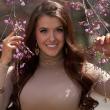 Photo of Maddie Duff, a young white woman with very long dark brown hair. She's wearing a cream colored turtle neck, make-up, and is smiling at the camera, She's standing among tree branches with small pink buds. 