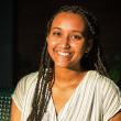 Snapshot of Segen Mussie, a young Black woman with long black twists. She's smiling, wearing a loose white, short-sleeved, v-neck blouse with thin gray vertical stripes. The background is dark but she seems to be sitting on a metal bench. 