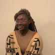 Photo of Treasure Ebikwo, a young Black Woman with long braids partially pulled back. She's wearing clear, plastic-rimmed glasses, a tan shirt, and a cream colored sweater with black and brown geometric lines. She's smiling and looking slight aside of the camera; behind her is a beige wall. 