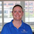 Headshot of Ellen Hartman, a white womane with blué eyes and dark hair pulled back tightly. She's smiling at the camera, wearing a blue polo shirt with the UK logo, and standing in front of a large window. 