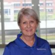 Headshot of Gail Sievert, a white woman with short gray hair. She's smiling at the camera, wearing a blue polo shirt and dangly blue earrings, and standing in front of a large window. 