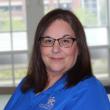 Headshot of Laura Ashe, a middle-aged white woman with dark straight hair just past her shoulders. She's smiling at the camera, wearing rectangular dark-rimmed glasses and a blue polo shirt, and standing in front of a large window. 