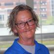 Haedshot of Terri Spear, a white woman with short, wavy gray hair and black plastic-rim glasses. She's smiling at the camera, wearing a blue polo shirt and standing in front a large window. 