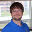 Headshot of Zachary Ross, a young white man with brown hair, a thin mustache and beard, and rectangular wire-framed glasses. He's smiling at the camera, wearing a blue polo shirt, and standing in front of a large window. 