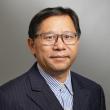 Headshot of Douglas Zhang, a middle-aged Asian man wearing a dark gray pinstripe suit with a light blue and white striped shirt. He has dark hair parted on his left side and plastic-rimmed glasses. He's giving a gentle smile. 