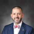 Headshot of Matt Bush, a white man with a buzz cut and short black and gray beard. He's smiling at the camera, wearing a blue suit jacket, red bow tie, and blue and red pocket square. A marbled gray backdrop is behind him. 