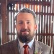 Headshot o Noah Perry, a white man with brown wavy hair pulled back and a brown mustache and beard. He's wearing a light brown plaid suit with a rust-colored tie. He's smiling at the camera, and behind him is a slatted wall through which sun is shining. 