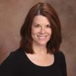 Professional headshot of Lynn Warneke, a white woman with straight, shoulder-length brown hair. She's wearing a black scoop-necked shirt and smiling at the camera. A brown photo backdrop is behind her. 