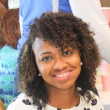 A young black woman with curly hair parted on the side and reaching just above her shoulders. She's smiling at the camera, wearing pearl stud earrings and a white blouse with black flowers. 