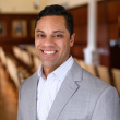 Professional photo of Antonio Garcia, a man with dark wavy hair parted on the side and brushed back. He's wearing a light gray suit and a white collared shirt that has a grid pattern of thin gray line. The background behind him is out of focus, a wood-paneled room with rows of upholstered chairs. 