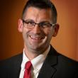 Professional studio headshot of Brian Noehren, a middle-aged white man with short dark hair, buzzed on sides and a bit longer on top. He's smiling at the camera, wearing rectangular glasses that are frameless on the bottoms of the lenses. He's in a dark gray suit, white shirt, and red tie. Behind him is a brownish-orange backdrop. 