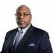 Professional studio photo of Darwin Conwell, a middle-aged Black man with buzzed gray hair and closely shaved mustache and beard. He's wearing a dark suit with a light blue tie and pocket square, and black rectangular glasses that are frameless on the bottom. 