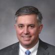 Professional studio headshot of Simon Fisher, a middle-aged white man with gray hair parted on the right side. He's wearing a light gray suit, white shirt, and light blue tie. He's smiling at the camera. Behind him is a marbled-gray backdrop. 