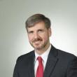 Professional studio photo of Kevin Hatton, a thin, middle-aged white man wearing a dark gray suit, white collared shirt, and red tie. He has light brown/gray hair parted on the side, and as short beard and mustache. He's smiling at the camera. 