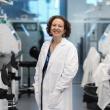 Professional photo of Ilhem Messaoudi, a middle-aged woman with brown, curly, chin-length hair. She's standing in a research lab, wearing a lab coat and smiling at the camera. 