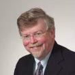 Professional studio headshot of John Fowlkes, a middle-aged white man with dark blonde hair parted on the side. He's wearing a dark gray suit, grayish purple satin tie, and rimless glasses. He has a dark blond mustache and goatee. 