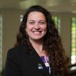 Professional photo of Luciana Shaddox, a young woman with long, curly, dark brown hair. She's smiling at the camera, wearing a dark suit jacket. in the background, out of focus, are large floor-to-ceiling windows. 