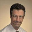 Professional studio headshot of Pedro Vera, a middle-aged man with short, dark curly hair, a faint mustache and goatee, and round wire-rimmed glasses. He's wearing a collared short and tie. Behind him is a beige backdrop. 