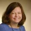 Professional studio headshot of Rebecca Dutch, a middle-aged white woman with brown hair to her shoulders. She's wearing a periwinkle shirt, a beaded necklace, and is smiling at the camera. Behind her is a tan backdrop. 