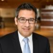 Professional headshot of Roshan Hussain, a middle-aged man with dark hair and dark-rimmed, rectangular glasses. He's wearing a dark gray suit, white shirt, and light blue tie. He's giving a slight smile to the camera. The out-of-focus background shows a wood-paneled room. 