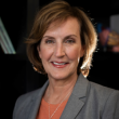 Professional headshot of Sharon Walsh, a white woman with chin-length brown hair. She's wearing a medium-gray suit jacket with a peach collared blouse. The background is dark, with a light shining in from the right side of the frame. 