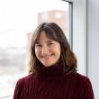 Photo of Jennifer Stevenson, a middle-aged white woman with shoulder length, wavy brown hair. She's smiling at the camera and wearing a dark red cable-knit turtleneck sweater. She's standing in front of a window. 