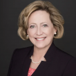 Professional headshot of a middle-aged white woman with dark blond hair cut just below her ears. She's wearing a black suit jacket, pink blouse, and pearl necklace with pearl earrings. Behind her is a dark gray backdrop. 