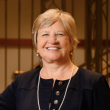 Photo of an older white woman with short blond hair. She's wearing a dark navy suit and long pearl necklack with pearl stud earrings. 