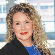 Photo of a white woman with long, curly blond hair. She's giving a calm smile to the camera and wearing red lipstick, a black blazer, and a royal blue shirt. Behind her are windows. 