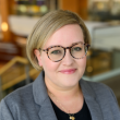 Professional headshot of a young white woman with a short blonde bob cut just below her ears. Shes's wearing tortoise-shell rimed glasses, a gray blazer, and dark shirt. She's smiling gently at the camera.  