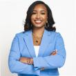 Professional photo of a young Black woman, smiling with her arms crossed. She has long black hair and is wearing a light blue suit with a white blouse. 