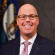 Man with a bald head and rectangular glasses smiles at camera wearing a dark blue suit, white shirt and light pink tie