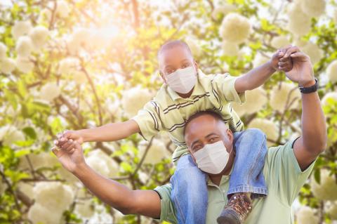 Black father with young son on his shoulder, both wearing masks. COVID