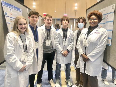 Six high school students in white coats stand between two research posters