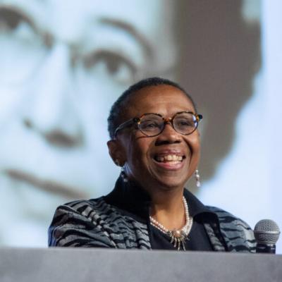 Photo of Vanessa Northington Gamble, a middle-aged Black woman with very short hair and glasses. She's standing a podium smiling, wearing a black and white blazer and on the screen behind her is the face of a woman. 