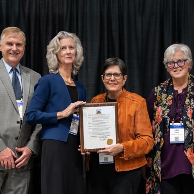 Photo of four people standing in front of dark stage curtains. Left to right: Philip Kern, Katherine Hartmann, Lexington Mayor Linda Gorton, and Lisa Cassis. Hartmann and Gorton are together holding a proclamation on a wooden plaque. 