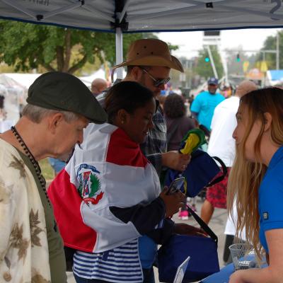 At a street festival, three UK staff members in blue polo shirts speak with three community members under canopy.