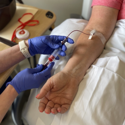 Two hands in blue medical gloves draw blood into a vial from someone's arm, which is resting on a pillow. 