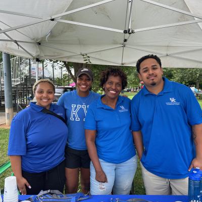 Four people in royal blue shirts stand together, smiling, under an event tent. They have Black and brown skin tones. There are bleachers in the background.